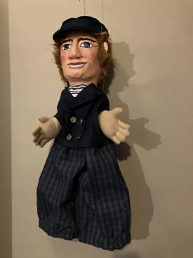 Homme vêtu d'un pantalon à carreaux, d'une veste noir, d'un pull à rayures et coiffé d'une casquette de pêcheur