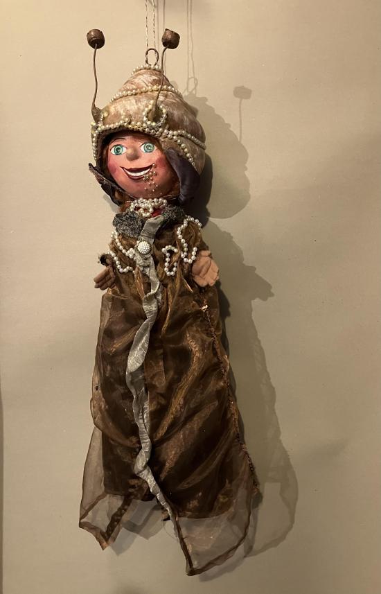 Escargote à tête de jeune fille, vêtue d'un habit brun, colliers de perles, elle porte sa coquille comme un chapeau